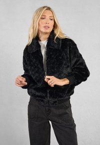 Giacca bomber in pelliccia sintetica nera con chiusura a zip, motivo a rombi e polsini a coste; indossata sopra una maglia chiara con jeans neri a vestibilità rilassata.