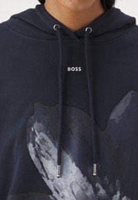 Sweat à capuche bleu marine avec cordons et embouts argentés, avec un design abstrait gris et le mot BOSS imprimé au-dessus de la poitrine.
