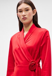 Czerwona sukienka w fasonie kopertowym, wykonana z gładkiego materiału, z długimi rękawami i dekoracyjnym akcentem w postaci róży w talii, podkreślająca swój dopasowany krój.