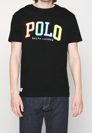 Osoba nosząca czarną koszulkę z kolorowymi literami "POLO" i napisem "Ralph Lauren", w ciemnoniebieskich dżinsach, stojąca z rozluźnionymi ramionami.