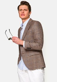 Jeune homme portant un blazer marron à carreaux, une chemise bleu clair rayée et un pantalon blanc, tenant des lunettes de soleil dans sa main droite.
