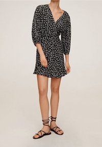 Schwarz-blumiges Wickelkleid mit langen Ärmeln, tailliertem Schnitt und ausgestelltem Rock, kombiniert mit braunen Sandalen mit Riemen. Strukturierter Stoff.