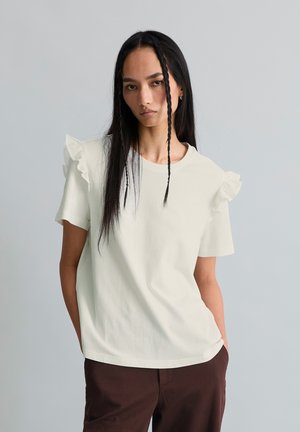 Femme aux longs cheveux noirs avec deux tresses à l'avant, portant un haut court à manches courtes à volants couleur blanc cassé et un pantalon foncé, debout devant un fond gris uni.