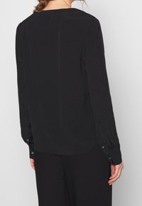 Blusa nera a maniche lunghe con un tessuto leggero e morbido, orlo curvo e polsini con bottone. Design semplice, senza motivi né accenti.