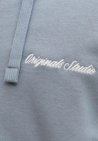 Sweat-shirt gris clair en maille texturée. Présente un logo brodé « Originals Studio » en blanc sur le devant, avec un détail à cordon de serrage.