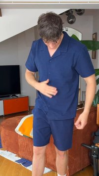 Άνδρας που φοράει ναυτικό μπλε κοντομάνικο πουκάμισο με κουμπιά και σορτς, προσαρμόζει το πουκάμισο σε ένα σαλόνι με τηλεόραση και πορτοκαλί καναπέ.