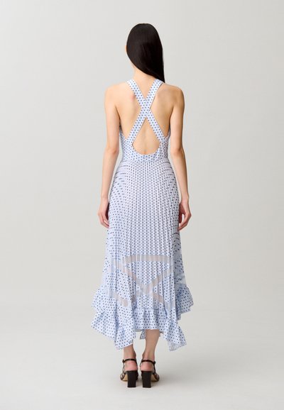 Donna con lunghi capelli scuri che indossa un vestito azzurro chiaro con pois, senza schienale, con spalline incrociate e orlo arricciato, in piedi davanti a uno sfondo semplice.