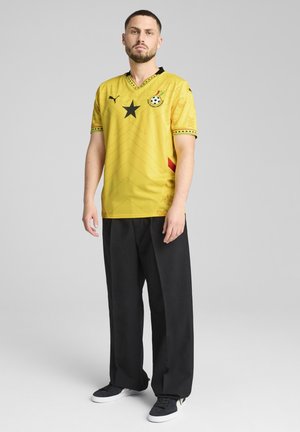 Tricou de fotbal galben cu accente negre, având un logo în formă de stea, design cu gât în V și modele geometrice. Asortat cu pantaloni negri și adidași.