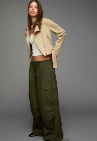 Beige mockasinskort jacka med dragkedja, vit åtsittande topp och oversize olivgröna cargobyxor med dragsko och fickor. Svarta skor.