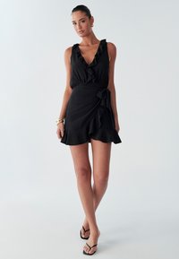 Schwarzes ärmelloses Kleid mit gerafftem Halsausschnitt und Wickeldesign, ausgestattet mit einem ausgestellten Saum und einem gebundenen Akzent an der Taille. Glatter Stoff.