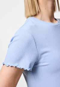 T-shirt à manches courtes de couleur bleu clair avec des côtes. Il présente un col rond et des bords de manches froncés, mettant en valeur une coupe ajustée et une texture douce.