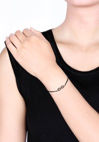 Bracelet en cordon noir arborant un symbole infini orné de petites pierres noires brillantes. Porté sur un poignet contre une chemise sombre.