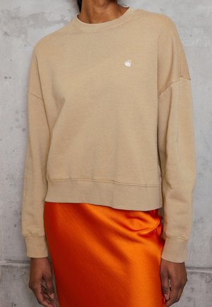 Osoba w beżowej, długorękawowej bluzie z małym logo i błyszczącej pomarańczowej spódnicy satynowej na tle szarej, teksturowanej ściany.