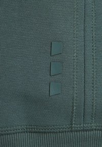 Un tessuto verde scuro testurizzato presenta un motivo a righe con tre accenti in silicone quadrati sul lato. Dettagli delle cuciture visibili.