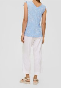 Donna che indossa un top senza maniche blu a fantasia, pantaloni bianchi a gamba larga e sandali con zeppa beige, in piedi di fronte a uno sfondo neutro.