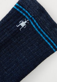 Tamsiai mėlyna kojinė su tekstūruotu audiniu, kurios viršuje yra šviesiai mėlynos juostos ir baltas figūros logotipas šalia krašto.
