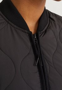 Giacca bomber nera trapuntata con collo a costine e zip frontale. Il materiale sembra liscio con motivi a onde testurizzate.