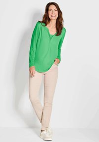 Grüne Langarmbluse mit V-Ausschnitt, kombiniert mit beigen, figurbetonten Hosen und weißen Sneakers. Glatter Stoff mit lockerem Schnitt.