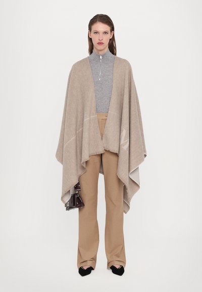 Beiger Poncho mit weicher Textur und umrahmenden Kanten, über einem grauen Reißverschluss-Oberteil und beigen Hosen getragen. Schwarze Absatzschuhe und eine dunkle Handtasche sind enthalten.