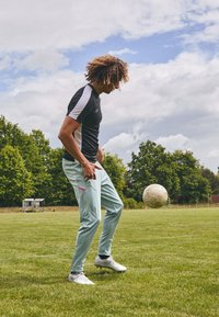 T-shirt de sport noir et blanc à manches courtes, jogging vert menthe clair avec des accents roses et chaussures de soccer blanches ; joueur contrôlant un ballon.