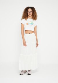 Witte crop top met kleurrijke palmprint, gecombineerd met een vloeiende witte maxi-rok met gelaagde tiers en een taille met trekkoord. Lichtgekleurde sneakers.