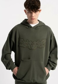 Oliwkowa, przewiewna bluza z kapturem o luźnym kroju, wykonana z fakturowanej tkaniny, z kieszenią kangurką i haftowanym wzorem na wysokości klatki piersiowej. Posiada sznurek do regulacji kaptura.