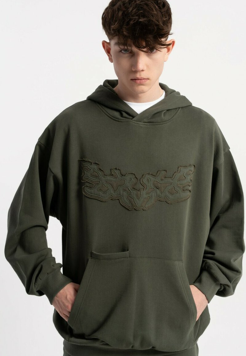 Oliwkowa, przewiewna bluza z kapturem o luźnym kroju, wykonana z fakturowanej tkaniny, z kieszenią kangurką i haftowanym wzorem na wysokości klatki piersiowej. Posiada sznurek do regulacji kaptura.