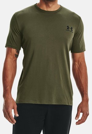 Mężczyzna w oliwkowozielonej koszulce z krótkim rękawem Under Armour i czarnych spodniach, z widocznym tatuażem na prawym przedramieniu.