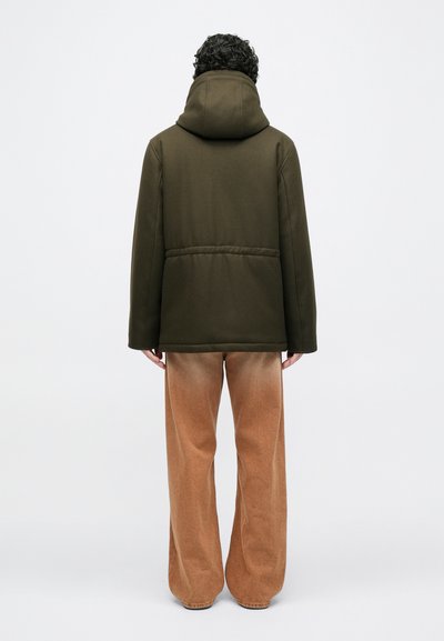 Oliivinvihreä hupullinen takki, jossa on sileä pinta ja takataskun sauma, yhdistettynä vaaleanruskeisiin leveälahkeisiin housuihin, jotka ovat hieman haalistuneet helmoista.