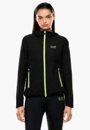 Donna che indossa una giacca nera con cappuccio e leggings con cerniere verde neon e loghi EA7 Emporio Armani, in piedi su sfondo bianco.