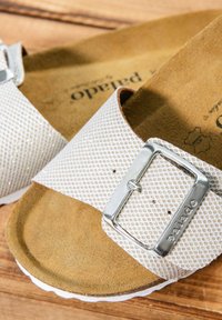 White perforated sandals with a metallic silver buckle, featuring a brown textured footbed and a white rubber sole.