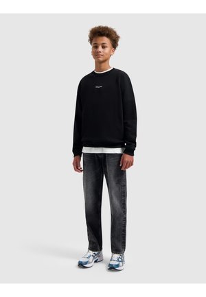 Tienerjongen staand, gekleed in een zwart sweatshirt, wit ondershirt, vervaagde zwarte spijkerbroek en wit-blauwe sneakers tegen een effen lichtgrijze achtergrond.