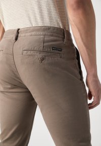 Braune Baumwollchinos mit glatter Textur, ausgestattet mit einer Knopftasche auf der R�ückseite und einem Logo-Label an der rechten hinteren Seite.