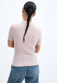 Vrouw met lang, gevlochten haar, gekleed in een lichtroze, gestructureerd T-shirt met korte mouwen en blauwe jeans, met gekruiste armen, met de rug naar de kijker gericht.