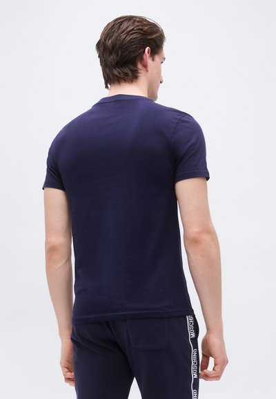 Homme portant un t-shirt à manches courtes bleu marine et un pantalon bleu marine avec des rayures blanches portant le logo "Moschino", debout de dos face au spectateur.