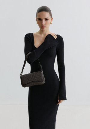 Kobieta z zaczesanymi do tyłu włosami, ubrana w czarną sukienkę z długim rękawem, trzymająca małą czarną plecioną torebkę na ramieniu.