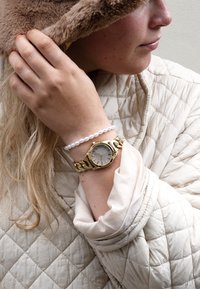 Montre-bracelet en or avec un cadran argenté texturé. Accompagnée d'un bracelet en perles blanches. Porte un manteau beige matelassé avec un col doux.