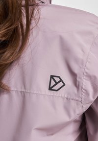 Heled lilla jakk, millel on sile tekstuur, musta geomeetrilise logo kaenla kohal. Reguleeritav krae ja puhtad õmblused.