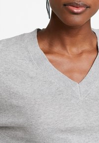 Šedý svetr s výstřihem do V z jemné pleteniny. Má žebrovaný límec a jemnou texturu. Detailní záběr zdůrazňuje límec a materiál.