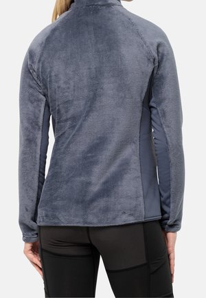 Femme portant une veste polaire grise ajustée et des leggings noirs, vue de dos sur un fond blanc uni.