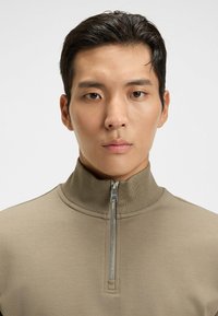 Quarter-zip pullover in beige fabric, featuring a high collar, smooth texture, and silver zipper detail at the neck.