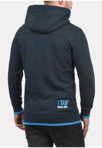 Marineblauer Kapuzenpullover aus weichem Stoff, mit hohem Kragen und einem kontrastierenden hellblauen Saum an den Bündchen und am Abschluss. Rückseite mit Logo und Text.