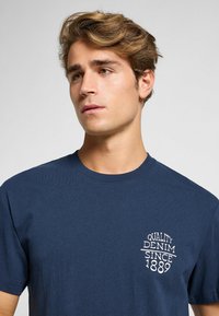 Camiseta de algodón azul marino con cuello redondo. Presenta el texto en blanco "Quality Denim Since 1889" en la parte izquierda del pecho. Textura suave.
