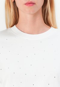 Bílá mikina s rozptýlenými malými stříbrnými a krystalovými ozdobami na přední straně, kulatým výstřihem a měkkou hladkou texturou látky.