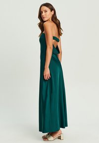 Lång, grön satin klänning med en strapless design, öppen rygg och volangdetalj på axlarna. Slät textur och flödande silhuett.