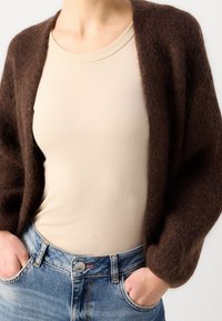 Braune, flauschige Strickjacke mit offener Vorderseite, getragen über einem beigefarbenen, figurbetonten Top und blauen Jeans. Die Strickjacke hat lange, weite Ärmel.