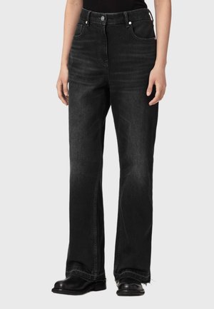 Person iført sorte jeans med lige ben med frynsete kanter og sorte lædersko, stående mod en ensfarvet baggrund.