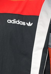 Τμήμα κόκκινου και μαύρου αθλητικού μπουφάν Adidas με λευκές ρίγες και κεντημένο λογότυπο Adidas και σύμβολο τριφύλλι.