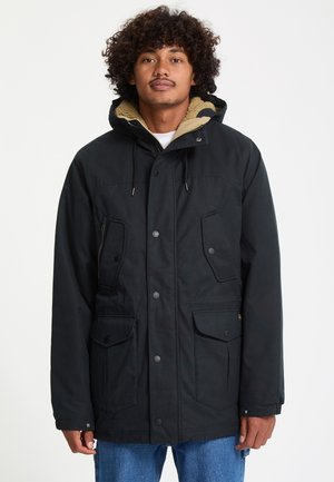 Homme aux cheveux bouclés portant un manteau d’hiver noir à capuche avec des poches avant et un jean bleu sur un fond blanc uni.