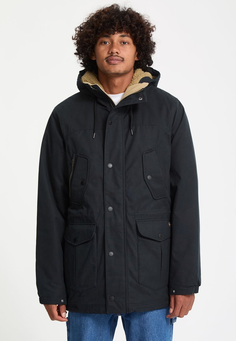 Homme aux cheveux bouclés portant un manteau d’hiver noir à capuche avec des poches avant et un jean bleu sur un fond blanc uni.